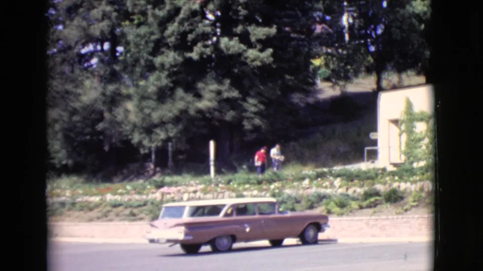 A tan station wagon passes a light-colored building as two distant pedestrians walk along the roadside in San Diego County.