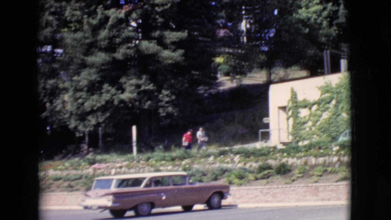 1968 San Diego County home-movie frame with a brown station wagon on the road and two men walking above a brick retaining wall.