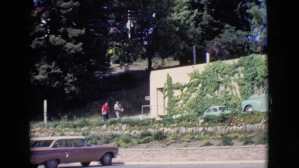 1968 San Diego County frame showing two men near a low ivy-covered building, with vintage cars visible around the landscaped roadside.