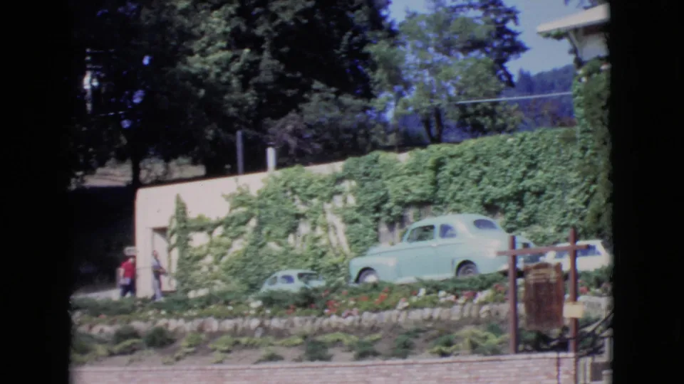 1968 San Diego County home-movie frame with two men near an ivy-covered building, a pale green vintage car, trees, and roadside foreground.