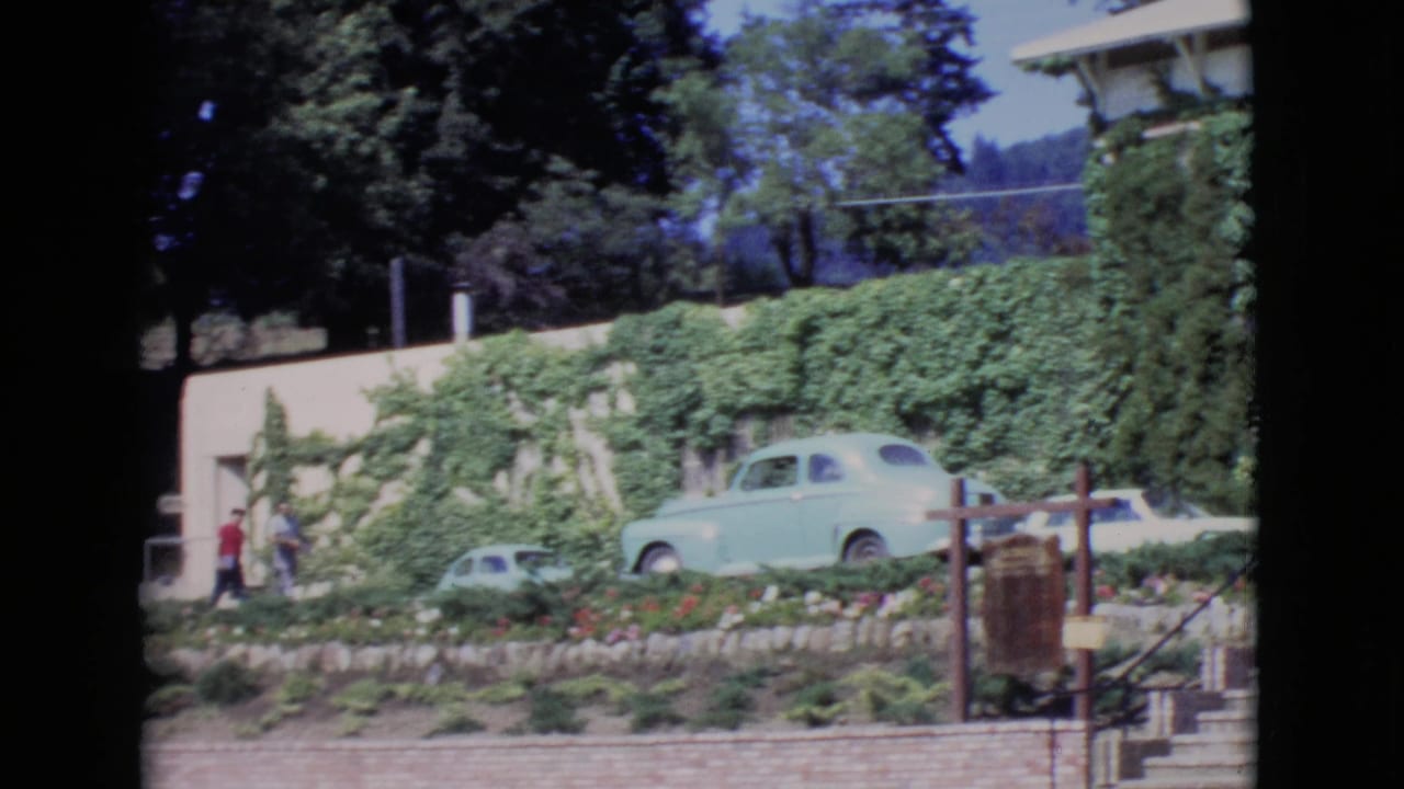 Ivy-covered building and garden with a light blue parked car and two people near the left side.