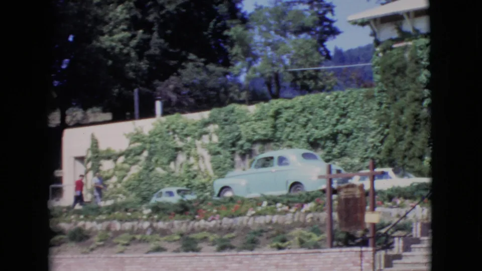 1968 San Diego County home-movie frame showing an ivy-covered exterior, garden beds, a light blue parked car, and two people at left.