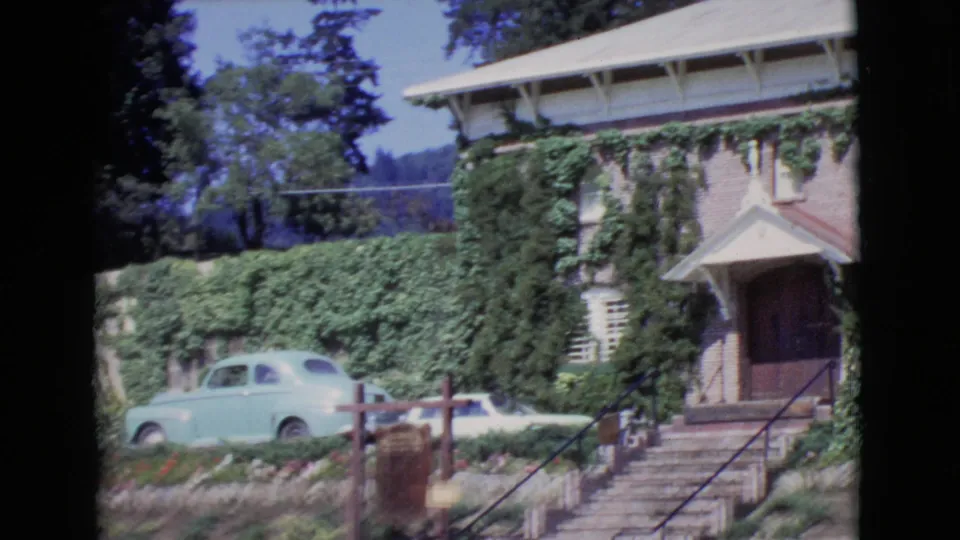 1968 San Diego County frame showing stairs to an ivy-covered building entrance with garden beds and a light blue car nearby.