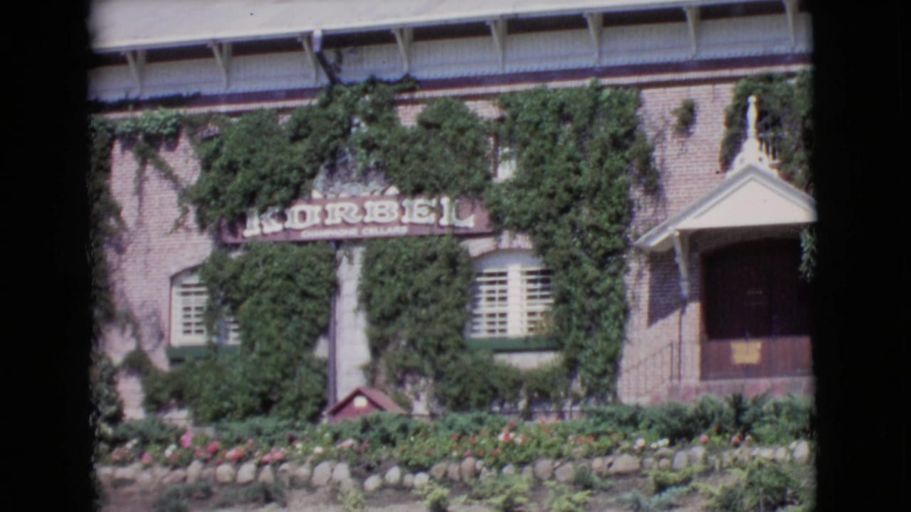 1968 San Diego County 8mm frame showing an ivy-covered brick facade with Korbel Champagne Cellars signage, garden flowers, and a small porch.