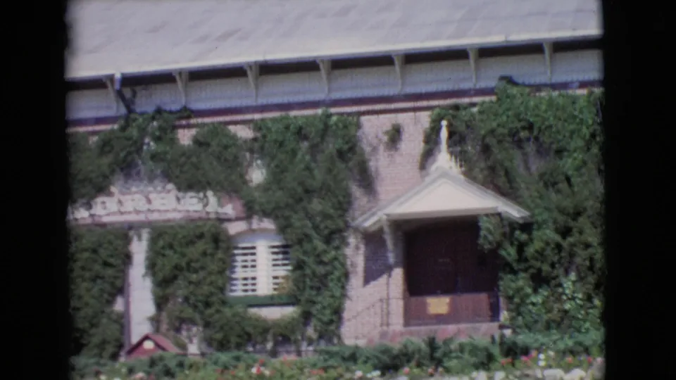 1968 San Diego County 8mm frame showing the lower facade, ivy, white shuttered windows, entrance porch, garden flowers, and stone edging.