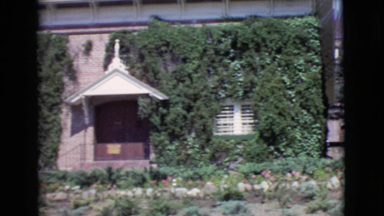 Ivy-covered brick building with gabled doorway, shuttered window, garden beds, and dark film-edge borders.