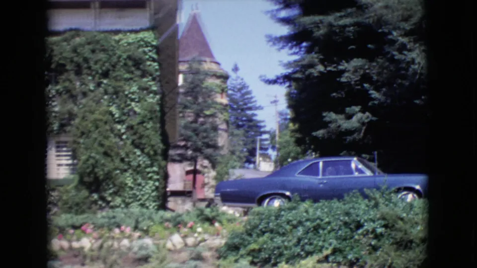 1968 San Diego County 8mm frame showing a stone tower with a conical roof beside an ivy-covered building and a blue parked car.
