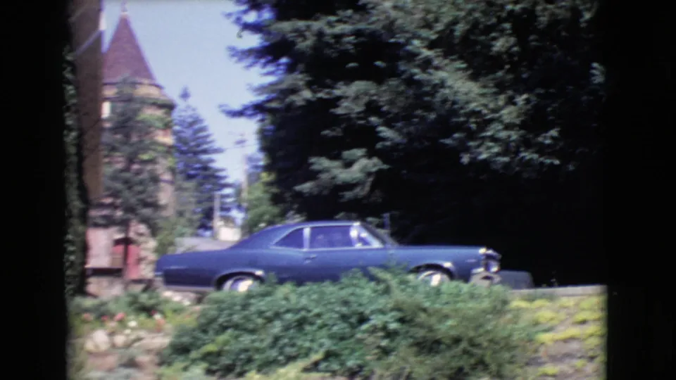 1968 San Diego County frame centered on a blue period car near dense shrubs, with a stone tower and conical roof behind it.
