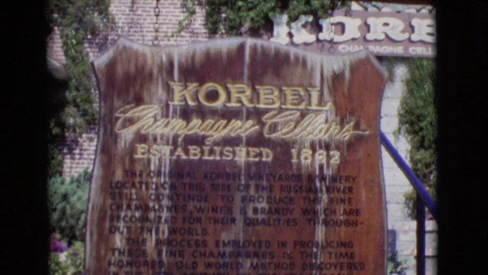 1968 8mm San Diego County frame showing a wooden Korbel Champagne Cellars sign, brick wall, ivy, steps, and blue handrail.