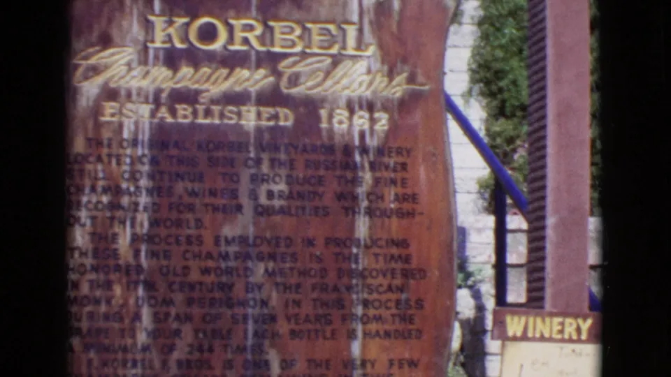 1968 8mm frame showing a weathered Korbel Champagne Cellars informational sign with gold lettering and a small winery sign nearby.
