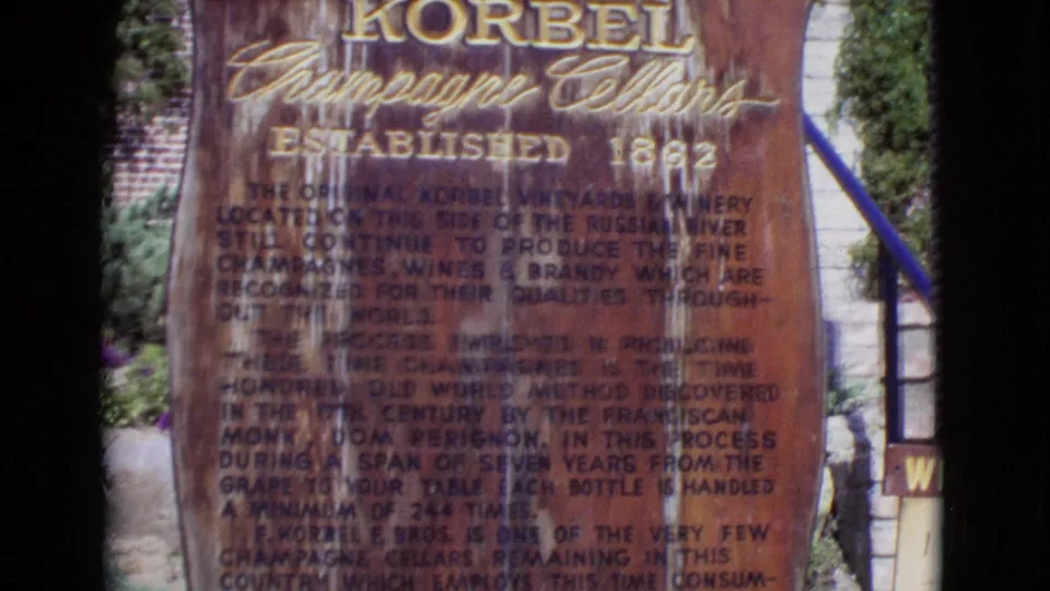 1968 California home-movie frame of the Korbel Champagne Cellars sign, with established date lettering, railing, stone wall, and foliage visible.