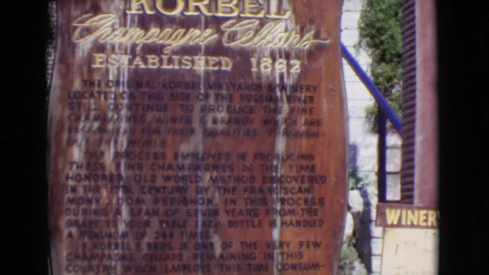 Close 8mm view of a weathered Korbel Champagne Cellars sign, with gold lettering and dense production-history text softened by film blur.