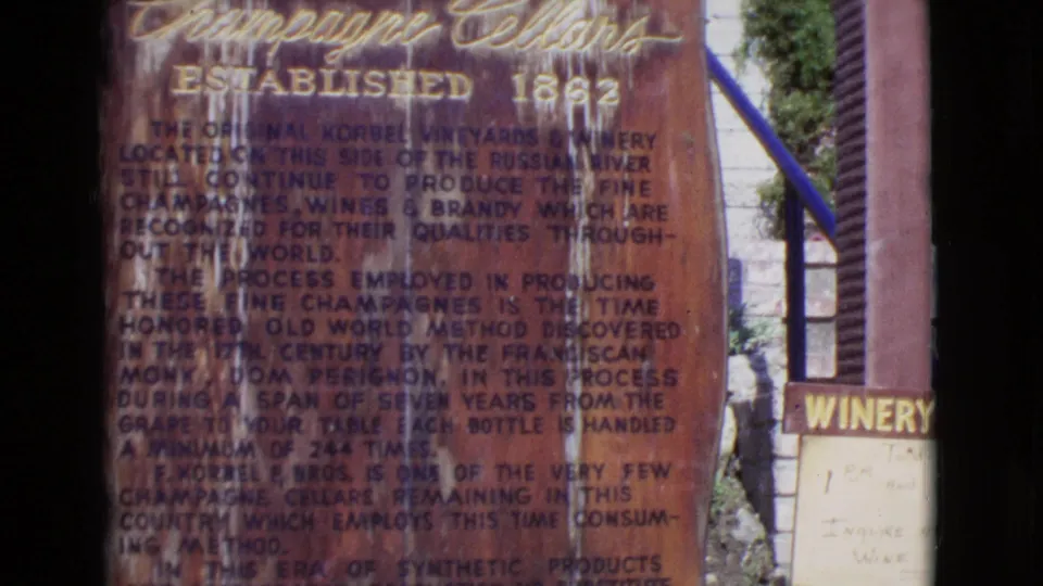 1968 8mm frame showing the Korbel Champagne Cellars informational sign beside a partial winery direction sign, railing, and garden foliage.