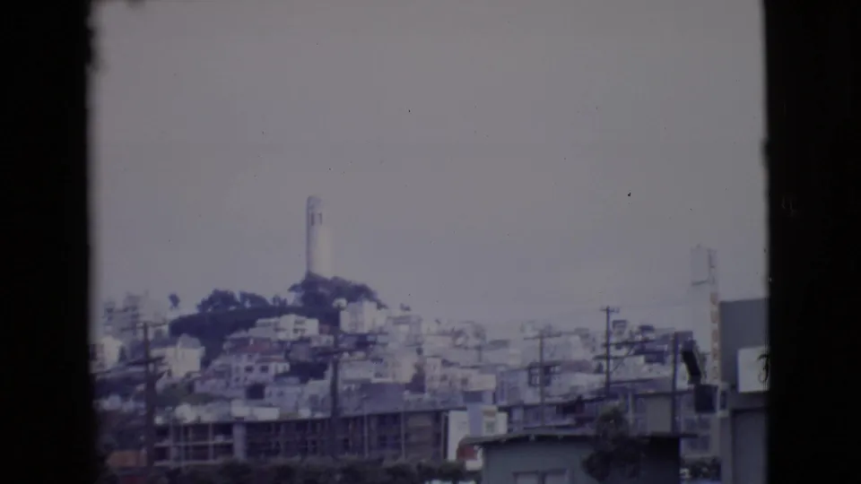 1968 San Diego County frame looking toward dense hillside buildings and a tall white tower beyond roadside power lines and low commercial structures.