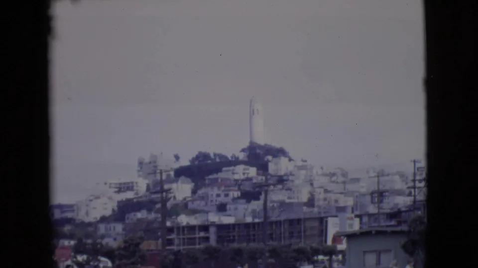 1968 San Diego County home-movie frame showing a wider hillside townscape with the white tower above clustered buildings and roadside utility poles.