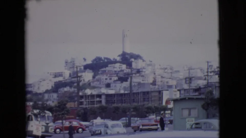 1968 home-movie frame showing a San Diego County hillside tower above street traffic, older cars, low buildings, and utility lines.