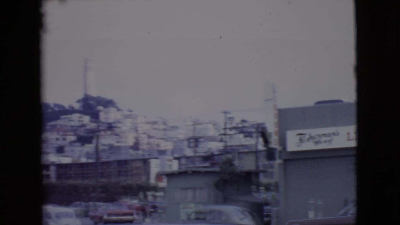 Soft 8mm view of a waterfront street with buildings, utility poles, distant cars, and dark film borders.