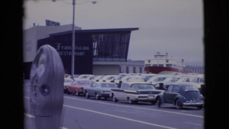 1968 San Diego frame showing a parking meter, classic cars, waterfront buildings, restaurant signage, and a ferry beyond the parking area.
