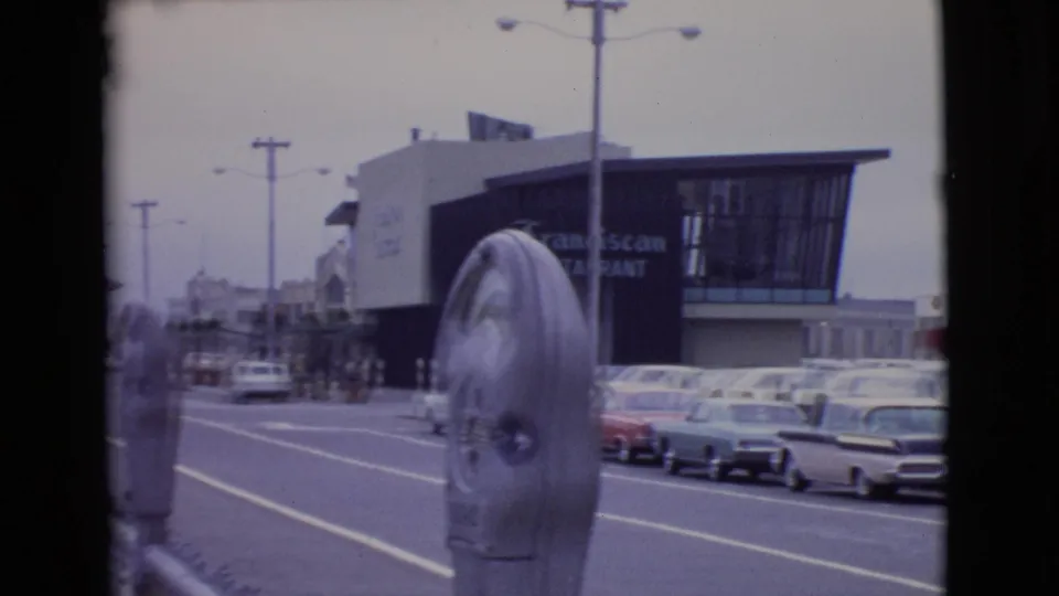 1968 San Diego home-movie frame looking past a parking meter toward parked cars, distant pedestrians, waterfront buildings, and restaurant signage.