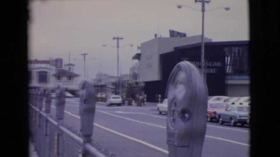 1968 San Diego County home-movie frame showing parking meters, roadway, parked cars, and storefront signage under an overcast sky.