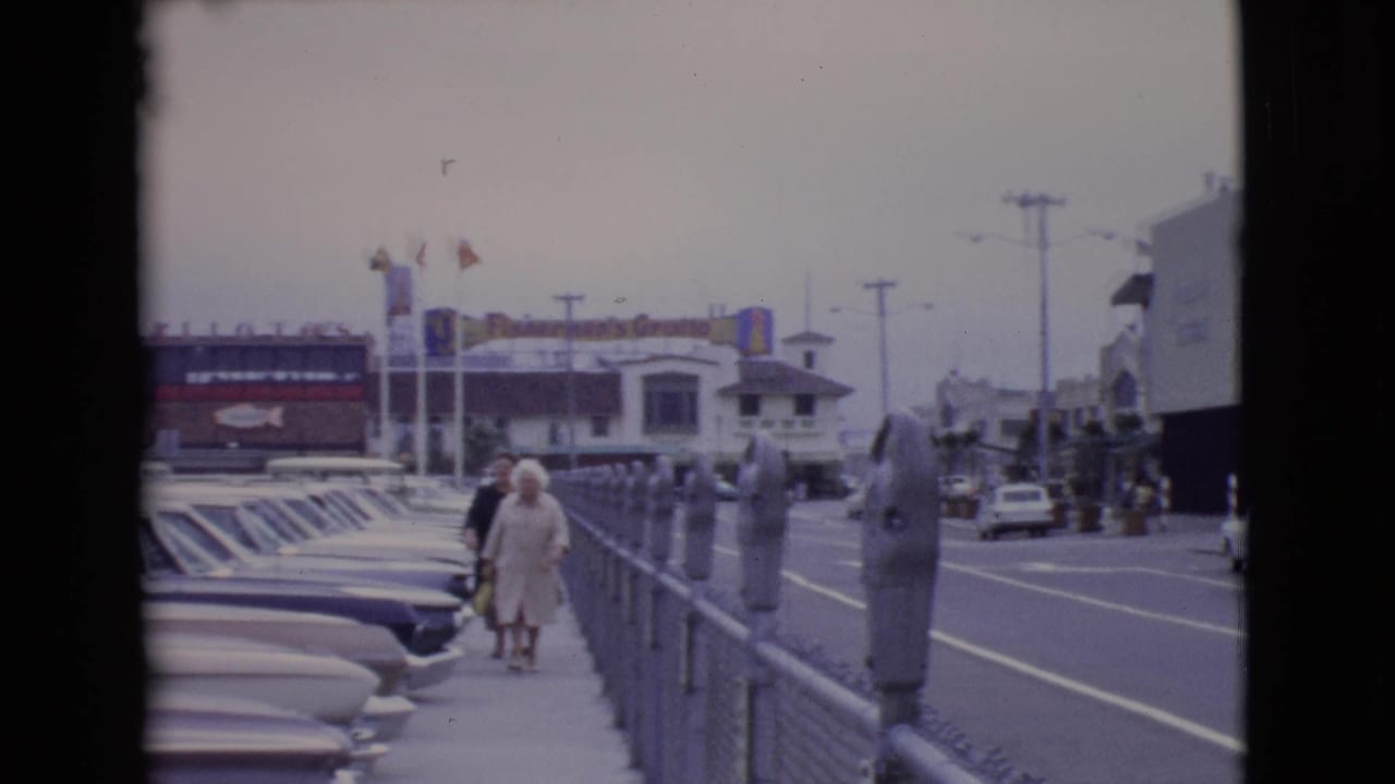 A couple walks along a San Diego County sidewalk in 1968, passing parking meters and rows of mid-century cars near visible business signs.