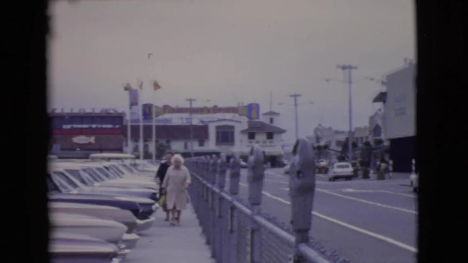 A couple walks along a San Diego County sidewalk in 1968, passing parking meters and rows of mid-century cars near visible business signs.