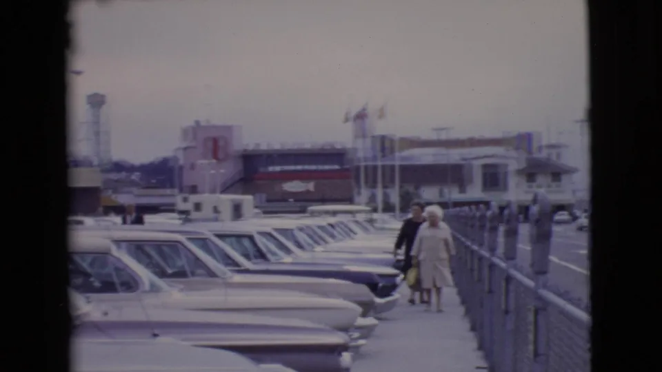 1968 San Diego County footage frame with a couple on the sidewalk, rows of parked cars, parking meters, flags, and storefront signage.