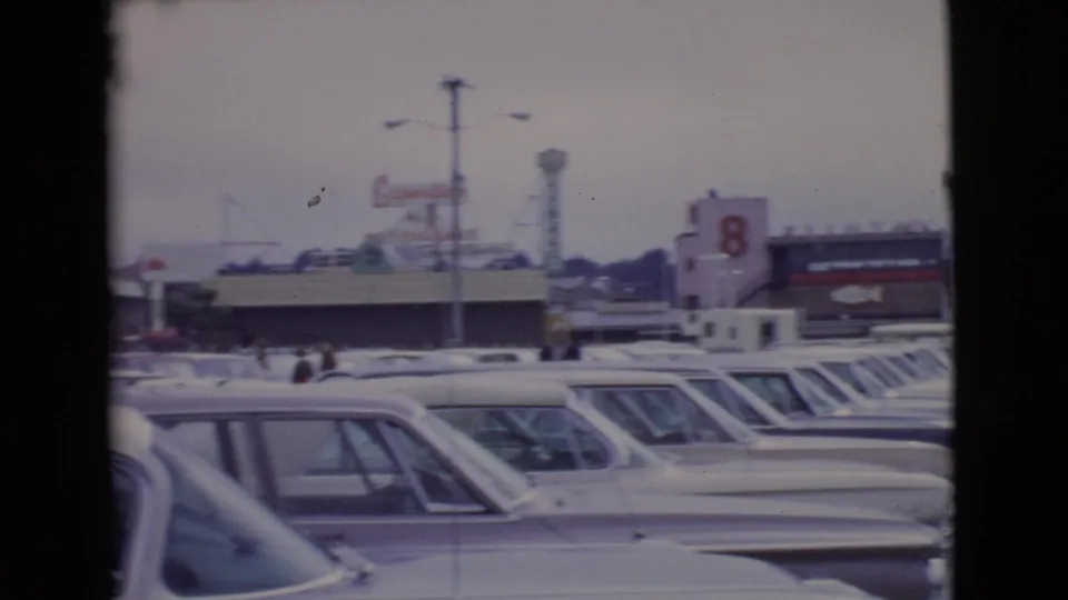 A 1968 San Diego County parking-lot view shows rows of cars, distant pedestrians, commercial buildings, and visible signs under gray light.