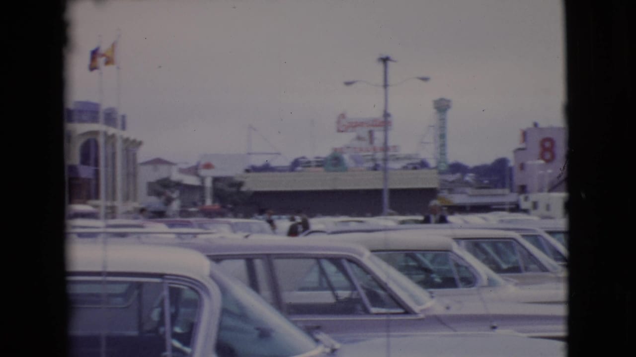 Crowded parking lot with 1960s cars near attraction buildings and flags.