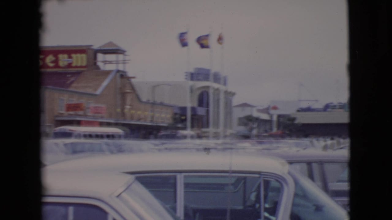 1968 San Diego County frame with wax museum signage, flags, a bus or van, and rows of parked cars in front.