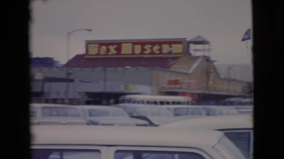 Motion-blurred 8mm frame in San Diego County showing wax museum signage, a bus, and mid-century cars during a handheld pan.