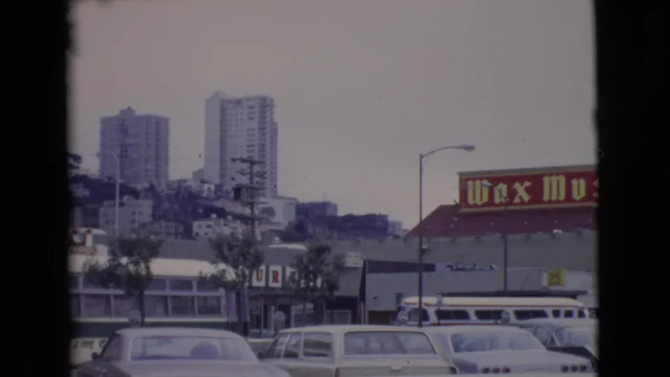 San Diego County frame showing parked 1960s cars, distant high-rise buildings, low storefronts, and part of the wax museum sign.