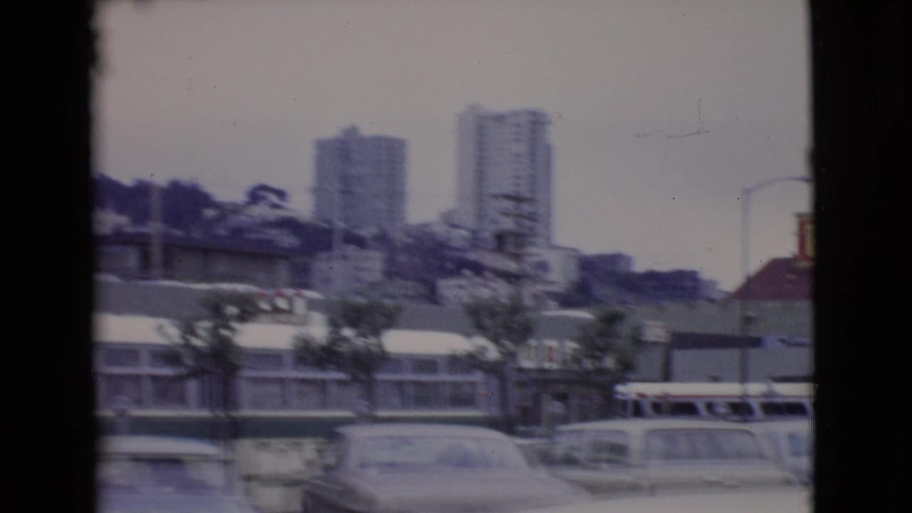 Soft 8mm street view with cars, buses, storefronts, and hillside apartment towers.