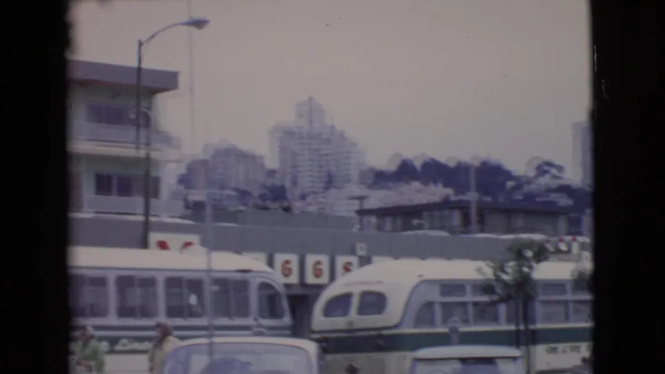 1968 San Diego County urban view with buses and cars in front of storefront signage, streetlights, and high-rise apartment buildings in the background.