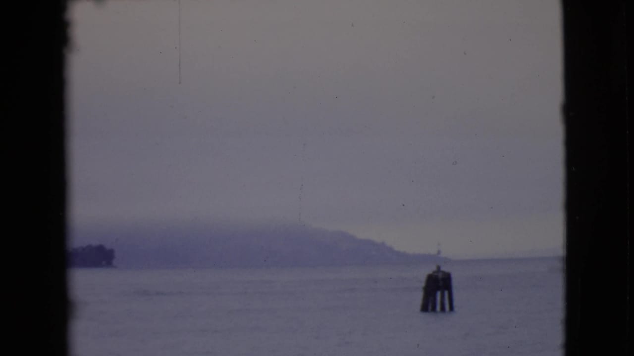 1968 8mm view across gray bay water toward a fog-softened island, with a channel marker in the foreground.