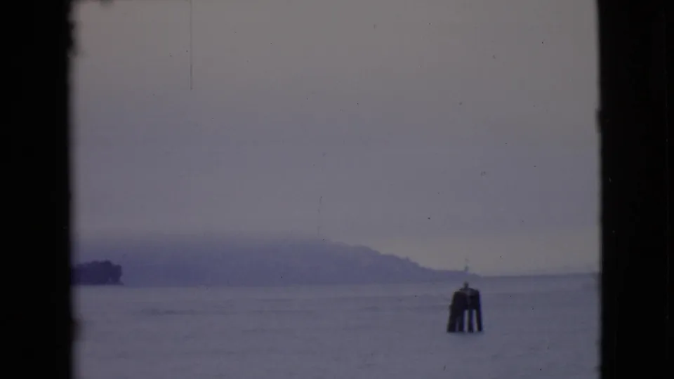 1968 8mm view across gray bay water toward a fog-softened island, with a channel marker in the foreground.