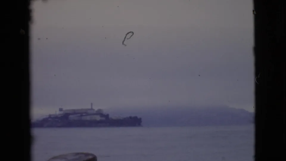 Fog-muted 1968 8mm view that appears to show Alcatraz Island across San Francisco Bay through a dark window-like frame.