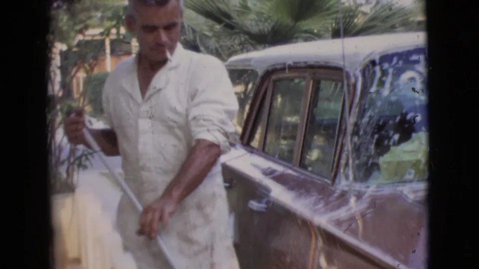 In San Diego County, 1968, an adult man in a white shirt washes a gold-colored classic car near palm greenery.