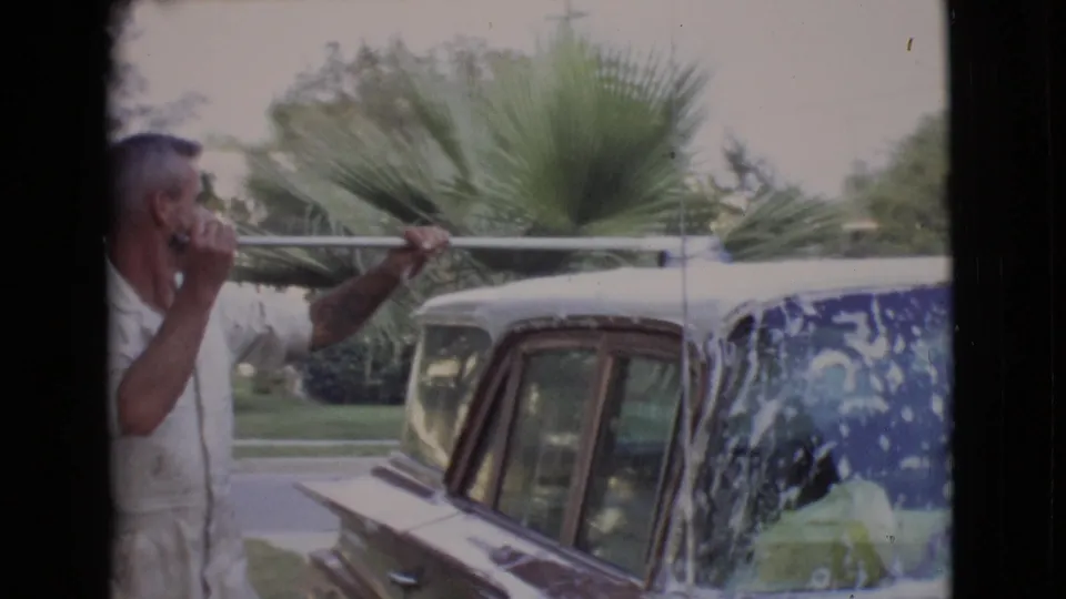 1968 San Diego County frame of an adult man scrubbing the roof area of a gold-colored classic car near palm plants.