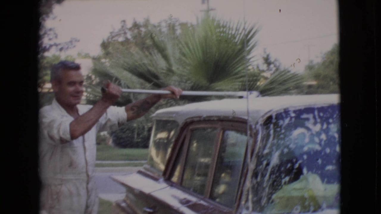 Man in white coveralls scrubs the roof of a soapy vintage car with a long-handled brush.