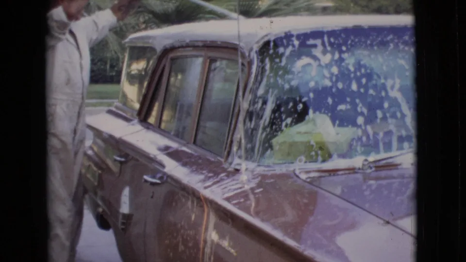 The 1968 home-movie frame shows the car's side and windshield covered with soap while the man washing it is partly cropped at left.