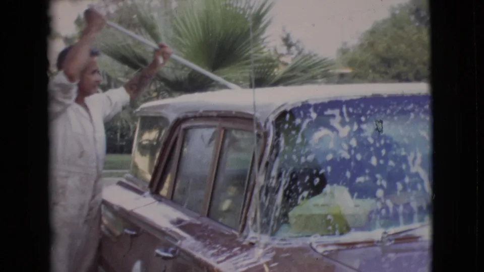 A man in white coveralls works the brush across a soapy vintage car in the driveway, with palm fronds visible behind him.