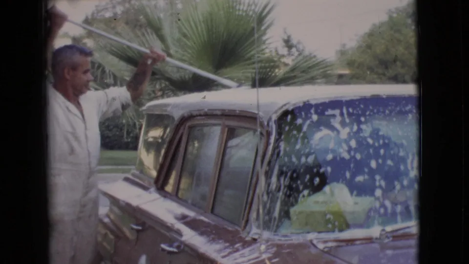 In a San Diego County driveway, the man continues brushing soap across the vintage car, with palm greenery and lawn behind the vehicle.