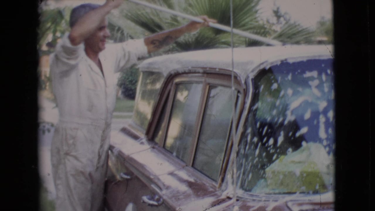 Man in white overalls raises a long-handled brush beside a soapy classic car.