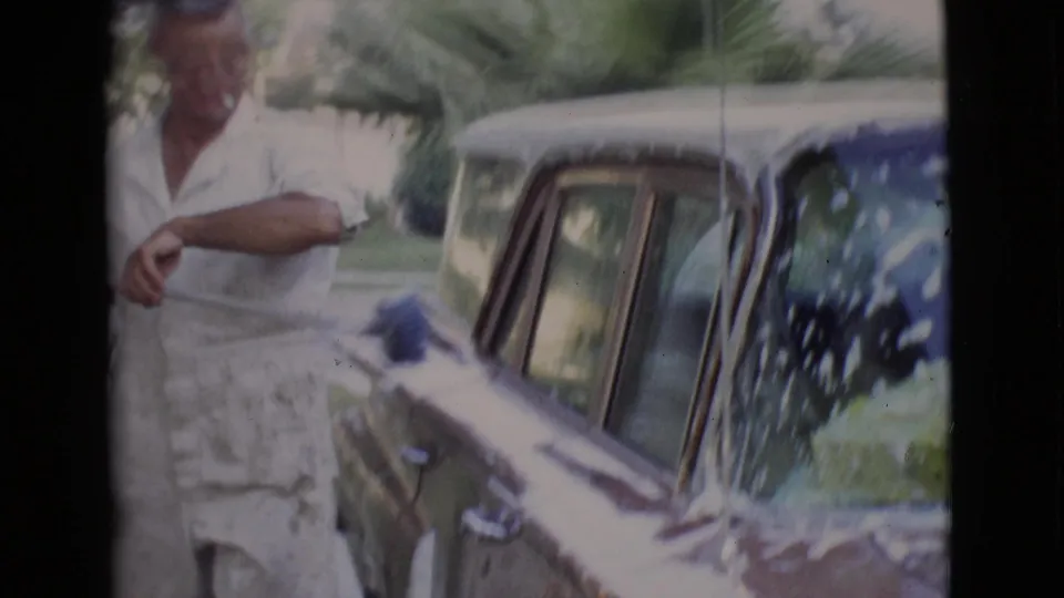 The man pauses next to the soapy car, holding the brush handle as the handheld 8mm frame catches the side windows and greenery.