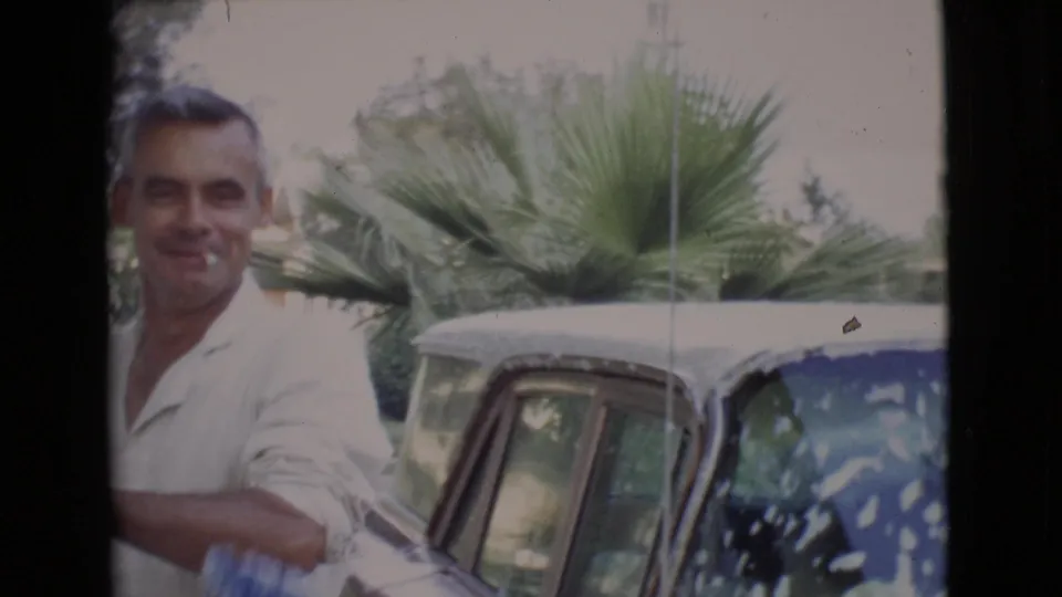The man smiles toward the camera with a cigarette visible while standing beside the soapy classic car in the 1968 San Diego County scene.