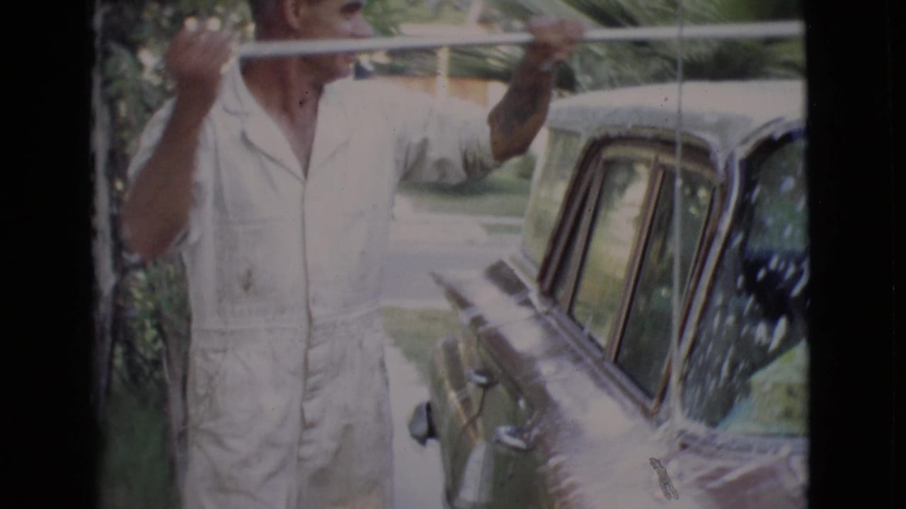 In 1968 San Diego County footage, a man in a light jumpsuit washes the roof of a classic car on a driveway.