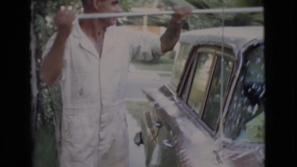 In 1968 San Diego County footage, a man in a light jumpsuit washes the roof of a classic car on a driveway.