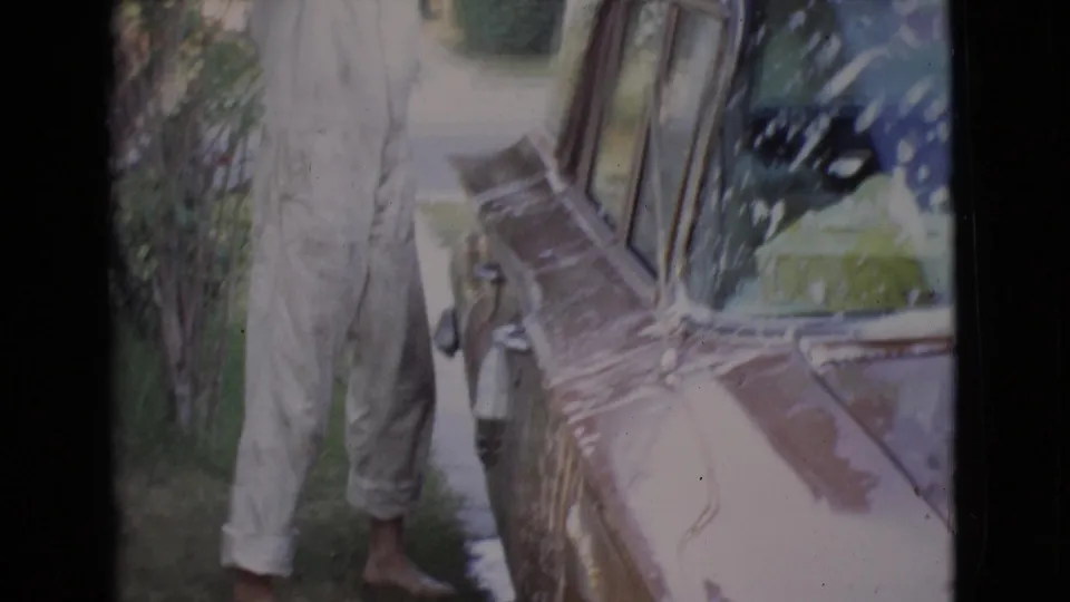 A man in a light jumpsuit stands by the soapy side of a classic car during a 1968 San Diego County home-movie scene.
