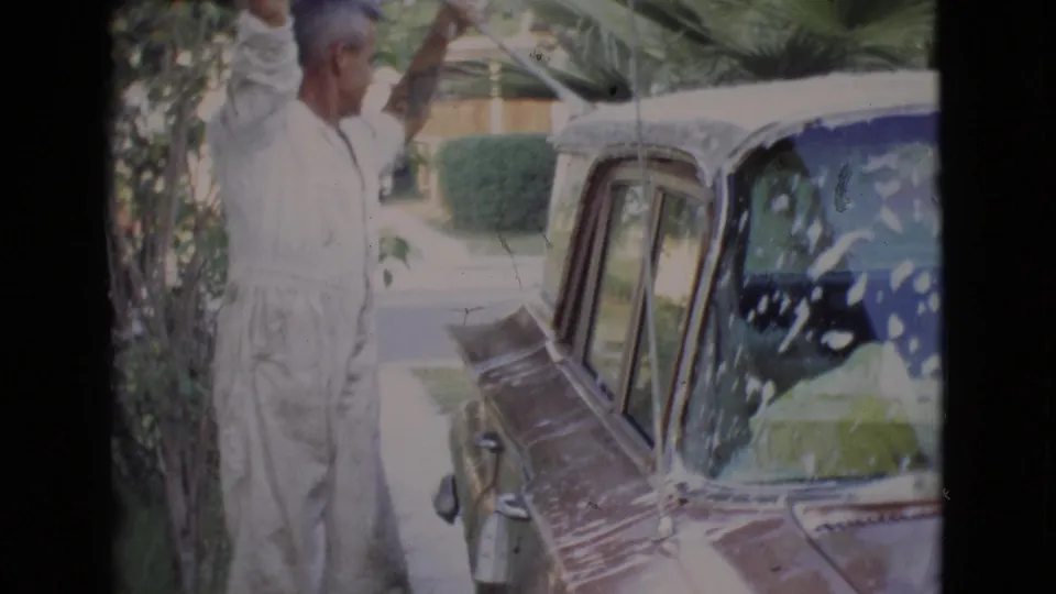 The man continues washing the soapy classic car beside shrubs and palm-like plants in San Diego County home-movie footage.